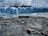 A large glacier with jagged icy peaks dominates the view. The glacier's blue hues contrast with the dark mountainous background. Foreground shows a rocky shoreline with a sign indicating the location as Parque Nacional Los Glaciares. Calm water reflects the glacier and the sky is overcast.