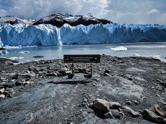 A large glacier with jagged icy peaks dominates the view. The glacier's blue hues contrast with the dark mountainous background. Foreground shows a rocky shoreline with a sign indicating the location as Parque Nacional Los Glaciares. Calm water reflects the glacier and the sky is overcast.