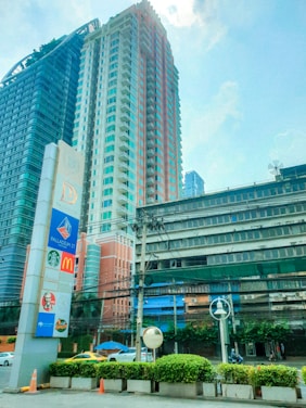 A tall modern building with a glass facade stands next to a slightly older structure with multiple windows. The foreground features a large vertical sign displaying logos of various well-known companies, including McDonald's and Starbucks. Trees and shrubs line the street below, and a yellow and green taxi is visible. The sky is blue with a few clouds.