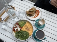 A bright, appetizing breakfast or brunch setting on a white wooden table. Includes a bowl with creamy scrambled eggs topped with avocado slices, sundried tomatoes with feta, and fresh arugula. A sandwich cut in half is plated with a decorative smear of orange sauce beside it. Two cups of coffee, one almost empty, are set in turquoise cups. A glass bottle of water and glasses accompany the meal.