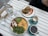 A bright, appetizing breakfast or brunch setting on a white wooden table. Includes a bowl with creamy scrambled eggs topped with avocado slices, sundried tomatoes with feta, and fresh arugula. A sandwich cut in half is plated with a decorative smear of orange sauce beside it. Two cups of coffee, one almost empty, are set in turquoise cups. A glass bottle of water and glasses accompany the meal.