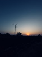 A wind turbine stands prominently against a dusky sky, accompanied by a setting sun on the horizon. The landscape is dotted with silhouettes of trees and power lines, creating a tranquil scene.