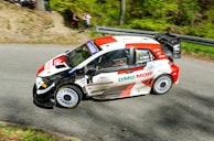 A rally car with a dynamic red, white, and black design speeds along a curving paved road. The vehicle features multiple sponsor logos and the driver’s and co-driver's names are visible on the side. Spectators can be seen in the background, partially obscured by foliage and trees, adding to the lively atmosphere.