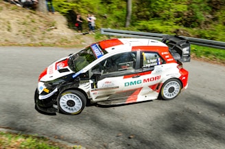 A rally car with a dynamic red, white, and black design speeds along a curving paved road. The vehicle features multiple sponsor logos and the driver’s and co-driver's names are visible on the side. Spectators can be seen in the background, partially obscured by foliage and trees, adding to the lively atmosphere.