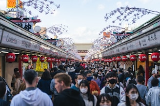 A bustling market street lined with shops, each adorned with vibrant red lanterns. The street is crowded with people, many wearing masks, under decorative branches. Signs in various languages are visible, showing a blend of cultures.