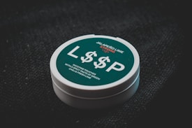 A container with a teal label rests on a dark textured surface. The label features the text 'Jalapeño Lime Strong 13mg', prominently displaying 'L$$P' in large white letters. Additional text mentions nicotine pouches with a spicy taste of green chili and fresh lime.