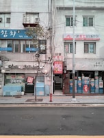 A street view of a row of small businesses in an urban area. The buildings have a mixture of signs in Korean, displaying a combination of blue, red, and white colors. A narrow sidewalk separates the storefronts from the street. Sparse trees and a streetlight are visible, adding to the urban atmosphere.