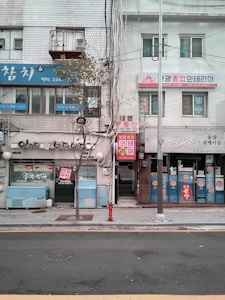 A street view of a row of small businesses in an urban area. The buildings have a mixture of signs in Korean, displaying a combination of blue, red, and white colors. A narrow sidewalk separates the storefronts from the street. Sparse trees and a streetlight are visible, adding to the urban atmosphere.