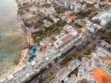 Aerial view of a coastal city featuring numerous multi-story buildings with rooftop pools, large residential complexes, and a mixture of modern and traditional architecture. The coastline has a beach with small boats docked near the shore. There are lush green areas interspersed among the buildings.