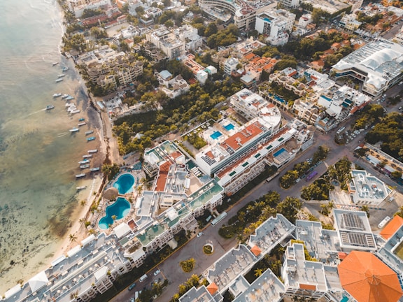 Aerial view of a coastal city featuring numerous multi-story buildings with rooftop pools, large residential complexes, and a mixture of modern and traditional architecture. The coastline has a beach with small boats docked near the shore. There are lush green areas interspersed among the buildings.