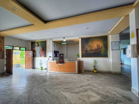 A spacious lobby features a reception desk at the center with two potted plants on either side. Above the desk, two large paintings are mounted on the walls; one depicts a serene figure with children, and the other illustrates a scene on a boat. The room has a gray marble floor and beige-colored ceiling details. A wooden door is partially open, allowing a view of the greenery outside.