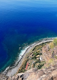 A breathtaking aerial view of a rugged coastline with a lush, green landscape below. The deep blue ocean dominates the upper portion, meeting a rocky shoreline where waves crash gently. The lower part features vibrant greenery and some structural elements, possibly buildings or agricultural setups nestled within the vegetation.