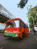 A bright orange and green bus is parked on the side of a street, with its door open. People are visible nearby on the sidewalk, and a tree with green leaves is overhead. An overpass runs parallel to the street.