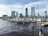 A city skyline with several modern high-rise buildings is visible in the background. In the foreground, a marina with multiple sailboats and yachts docked along wooden piers extends across a body of water. The water reflects the afternoon sunlight, creating a serene atmosphere. The sky is partly cloudy with a blue tint.