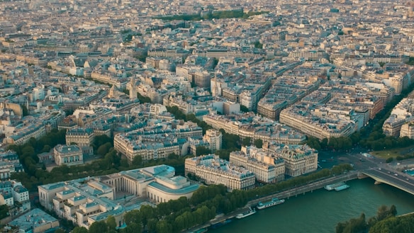 A large aerial view of a city with dense buildings and intricate streets. The image shows a notable concentration of architectural structures including notable landmarks, residential and commercial buildings. A river cuts through the bottom right of the image with bridges connecting the two sides of the city. Green spaces are scattered throughout, adding a touch of nature to the urban landscape.