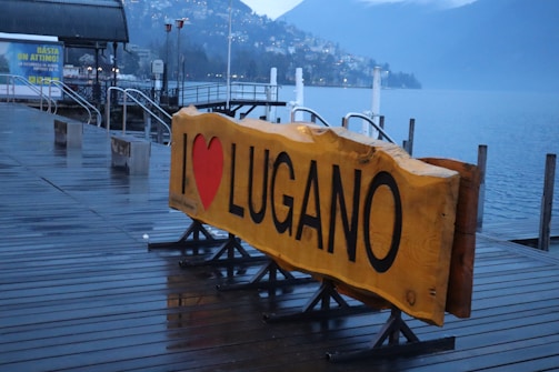A large wooden sign with bold black letters on a yellow background reads 'I ♥ LUGANO'. It is positioned on a wet wooden deck next to a serene lake with surrounding mountains partially obscured by mist. Streetlamps and a distant view of a town are visible, emphasizing a tranquil, scenic waterfront setting.