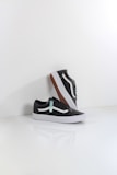 A pair of black sneakers with white soles and a distinctive white side stripe, set against a minimalistic white background. One shoe is positioned upright, resting against the other, showcasing the brown waffle-patterned outsole.