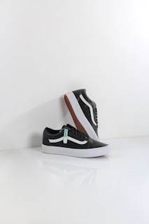 A pair of black sneakers with white soles and a distinctive white side stripe, set against a minimalistic white background. One shoe is positioned upright, resting against the other, showcasing the brown waffle-patterned outsole.
