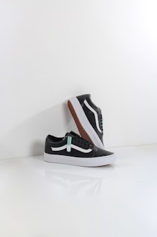 A pair of black sneakers with white soles and a distinctive white side stripe, set against a minimalistic white background. One shoe is positioned upright, resting against the other, showcasing the brown waffle-patterned outsole.
