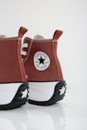 A pair of brown high-top Converse sneakers with distinctive thick white and black soles. The shoes feature the iconic Converse All Star logo on the heel and have white stitching and laces.