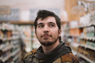 A young man with a beard and short hair stands in an aisle of a store, appearing thoughtful and slightly looking upwards. The background is blurred, showing shelves filled with various products.