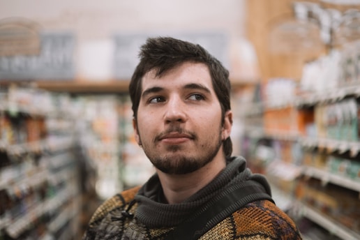 A young man with a beard and short hair stands in an aisle of a store, appearing thoughtful and slightly looking upwards. The background is blurred, showing shelves filled with various products.