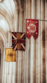 A set of three ceremonial flags displayed in a historical or religious building with tall, arched stone columns. The flags feature intricate designs with regal symbols, predominantly in red, blue, and yellow.