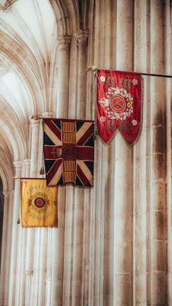 A set of three ceremonial flags displayed in a historical or religious building with tall, arched stone columns. The flags feature intricate designs with regal symbols, predominantly in red, blue, and yellow.