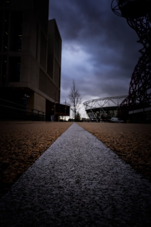 A perspective view of a pathway with a white line running down its center, emphasizing depth towards the horizon. Large buildings and a ferris wheel-like structure frame the sides under a gloomy, overcast sky.