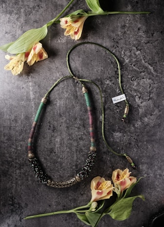 A handcrafted necklace with intricate beadwork and metal detailing lies on a textured stone surface. Surrounding the necklace are delicate yellow and pink flowers with long green stems and leaves. The overall appearance is both rustic and elegant, blending natural elements with artisan jewelry.