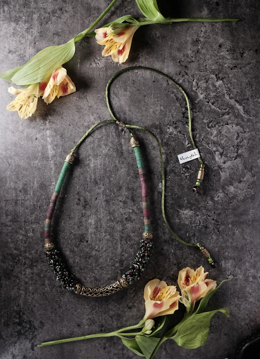 Close-up of handcrafted jewelry with warm pink and purple tones surrounded by delicate flowers and green leaves.