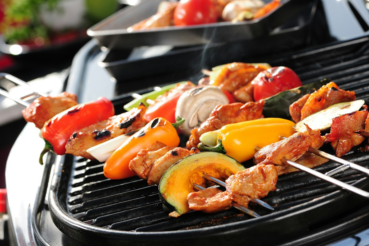 Brochetas de pollo, morrones, zapallo y champiñones dorándose sobre la placa estriada negra de una parrilla compacta, con una bandeja de verduras al fondo