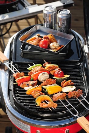 A barbecue grill cooking skewers of assorted vegetables and meat, with colorful bell peppers, mushrooms, and zucchini. Two beverage cans are placed on the side, and a tray with more grilled vegetables is nearby.