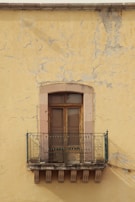 A rustic balcony with ornate metal railings is attached to an aged, weathered yellow wall. The wall has visible cracks and patches of peeling paint, giving it a worn, historical appearance. The balcony features wooden double doors with a stone frame, adding to the vintage charm.