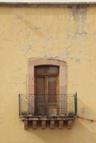 A rustic balcony with ornate metal railings is attached to an aged, weathered yellow wall. The wall has visible cracks and patches of peeling paint, giving it a worn, historical appearance. The balcony features wooden double doors with a stone frame, adding to the vintage charm.
