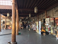A bustling marketplace with wooden columns and a variety of shops displaying colorful goods. People are casually strolling and shopping, while one person sits on a stool examining something in their hands. The scene is adorned with traditional lanterns hanging from the ceiling and the area has a traditional architectural charm.