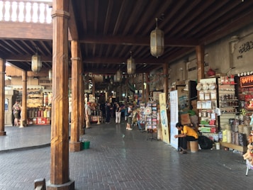 A bustling marketplace with wooden columns and a variety of shops displaying colorful goods. People are casually strolling and shopping, while one person sits on a stool examining something in their hands. The scene is adorned with traditional lanterns hanging from the ceiling and the area has a traditional architectural charm.