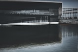 A modern architectural structure features clean lines and geometric shapes, reflecting in the still water below. The building is constructed with dark, sleek materials, creating a sense of minimalism and sophistication. Water cascades gently down the building into the pool, adding a dynamic element to the otherwise still scene.