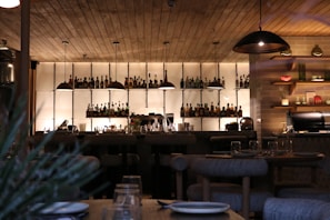 A dimly lit bar with wooden ceilings and shelves lined with various bottles. Hanging pendant lights illuminate the bar top, which is set with glassware and plates. The atmosphere appears cozy and intimate, typical of a modern restaurant or lounge area. To the right, there are shelves with decorative items and a kitchen area.