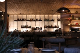 A dimly lit bar with wooden ceilings and shelves lined with various bottles. Hanging pendant lights illuminate the bar top, which is set with glassware and plates. The atmosphere appears cozy and intimate, typical of a modern restaurant or lounge area. To the right, there are shelves with decorative items and a kitchen area.
