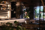 A modern indoor restaurant setting with neatly arranged tables and chairs. The bar area features a well-organized array of bottles on shelves against a striped black and white background. Potted plants and elegant lighting enhance the ambient atmosphere, with large windows allowing natural light to illuminate the room.