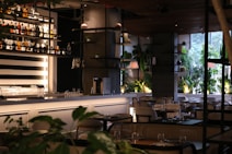A modern indoor restaurant setting with neatly arranged tables and chairs. The bar area features a well-organized array of bottles on shelves against a striped black and white background. Potted plants and elegant lighting enhance the ambient atmosphere, with large windows allowing natural light to illuminate the room.