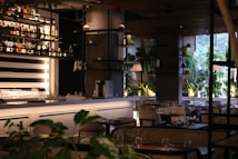 A modern indoor restaurant setting with neatly arranged tables and chairs. The bar area features a well-organized array of bottles on shelves against a striped black and white background. Potted plants and elegant lighting enhance the ambient atmosphere, with large windows allowing natural light to illuminate the room.