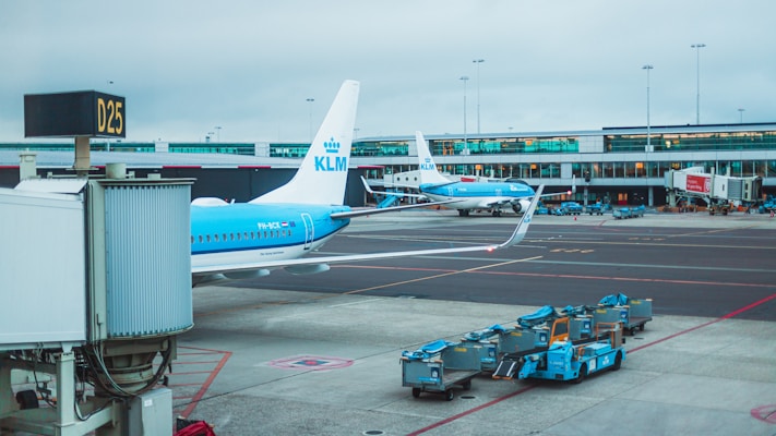 A busy airport scene featuring two airplanes on the tarmac, marked with KLM branding. In the foreground, ground service equipment and carts stand nearby. The airport terminal is visible in the background, housing multiple gates and large windows. The weather appears overcast, casting a muted light over the scene.