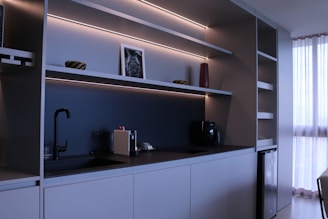 A modern kitchen with minimalist design featuring sleek cabinets, open shelves with LED lighting, a black faucet, and a coffee machine. A small framed picture, a red vase, and two decorative bowls are placed on the shelves. To the right, sunlight filters through white curtains, offering a sense of natural light.