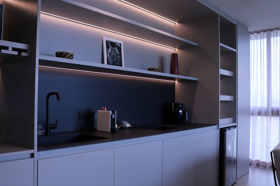 A modern kitchen with minimalist design featuring sleek cabinets, open shelves with LED lighting, a black faucet, and a coffee machine. A small framed picture, a red vase, and two decorative bowls are placed on the shelves. To the right, sunlight filters through white curtains, offering a sense of natural light.