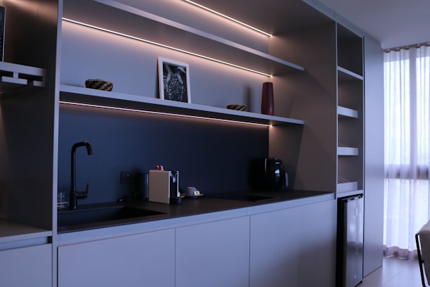 A modern kitchen with minimalist design featuring sleek cabinets, open shelves with LED lighting, a black faucet, and a coffee machine. A small framed picture, a red vase, and two decorative bowls are placed on the shelves. To the right, sunlight filters through white curtains, offering a sense of natural light.