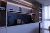 A modern kitchen with minimalist design featuring sleek cabinets, open shelves with LED lighting, a black faucet, and a coffee machine. A small framed picture, a red vase, and two decorative bowls are placed on the shelves. To the right, sunlight filters through white curtains, offering a sense of natural light.