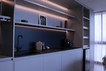 A modern kitchen with minimalist design featuring sleek cabinets, open shelves with LED lighting, a black faucet, and a coffee machine. A small framed picture, a red vase, and two decorative bowls are placed on the shelves. To the right, sunlight filters through white curtains, offering a sense of natural light.
