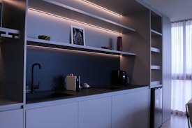A modern kitchen with minimalist design featuring sleek cabinets, open shelves with LED lighting, a black faucet, and a coffee machine. A small framed picture, a red vase, and two decorative bowls are placed on the shelves. To the right, sunlight filters through white curtains, offering a sense of natural light.